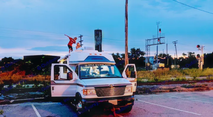 RV rental fun at dusk: Kids on a camper van roof by railroad tracks, one dabbing, others inside. Adventure awaits!. Chinook Concourse 2001
