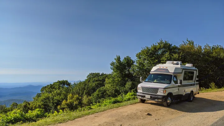 Scenic RV rental adventure: White camper van parked on a dirt road overlooking green mountains under a clear blue sky.. Chinook Concourse 2001