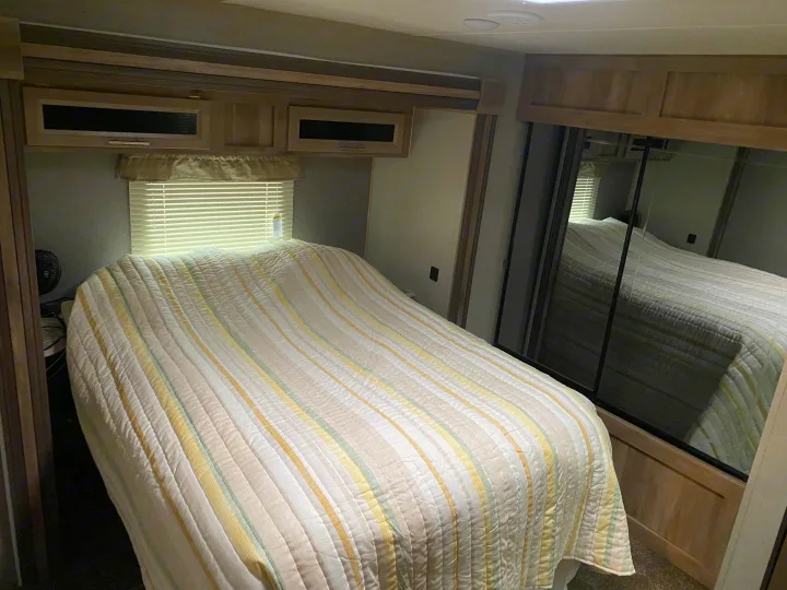 Inside this RV rental, a bright bedroom features a striped bedspread, window, overhead cabinets, and a mirrored wardrobe.. Coachmen Catalina 2021