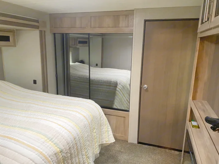 This RV rental bedroom features a striped bed, large mirrored closet, wooden door, and light wood cabinetry for a compact interior.. Coachmen Catalina 2021