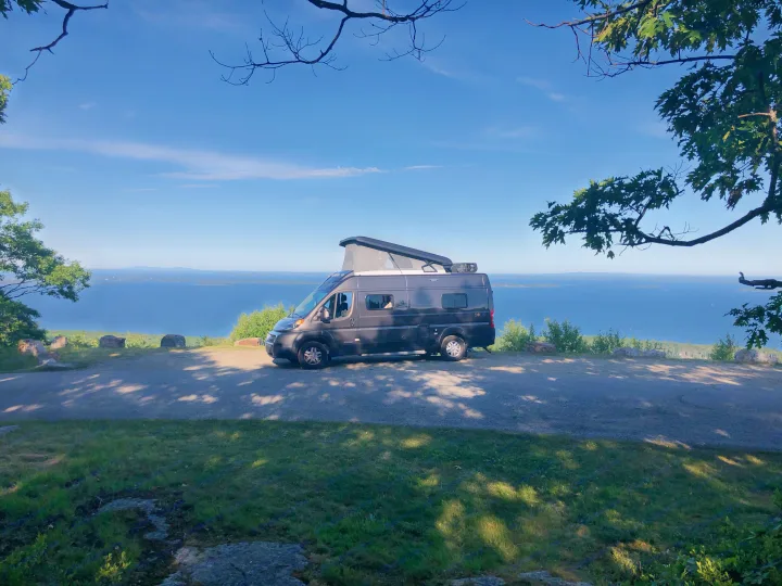 A dark gray RV rental with its pop-top open is parked at a scenic coastal overlook, offering stunning blue ocean and island views.. Winnebago Solis 2022