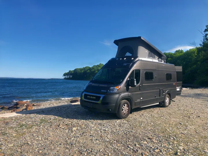 Dark grey RV rental camper van on a rocky beach by the ocean. Its pop-top is raised, ready for adventure under a clear blue sky.. Winnebago Solis 2022