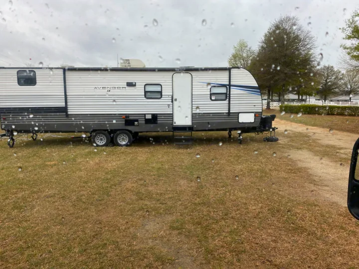 An Avenger LE travel trailer in a grassy lot, viewed through a rain-splashed window. Ideal RV rental for your next adventure.. Forest River All American Sport 2020