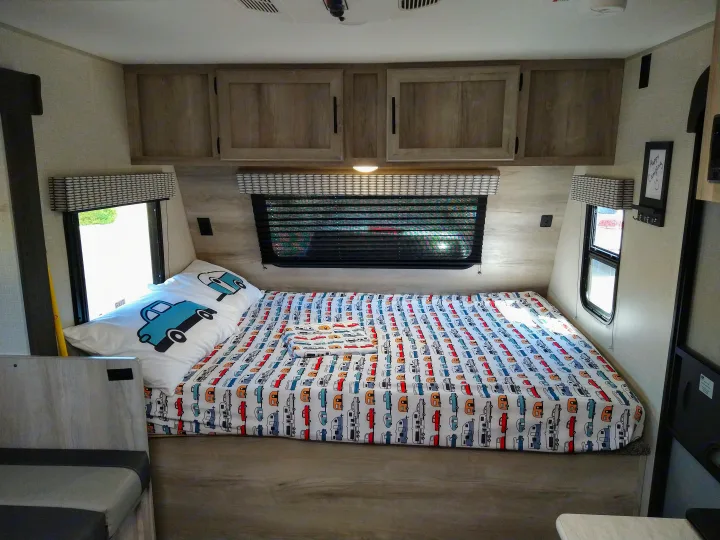 Cozy RV rental bed with blue truck pillow & playful camper-patterned sheets. Bright, functional interior.. KZ 181BH 2021
