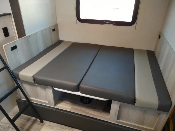 An RV rental interior showing a versatile bunk bed/dinette with grey cushions, built-in storage, and an emergency exit window.. KZ 181BH 2021
