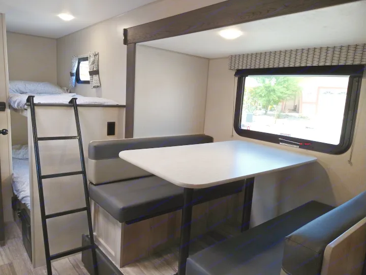 RV rental interior with bunk beds, a ladder, and a dinette area with table and benches. A window shows the outside.. KZ 181BH 2021