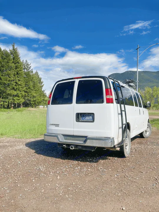 White RV rental van on a gravel road, surrounded by green trees, mountains, and a bright blue sky. Perfect for adventures.. Chevrolet Express 2007