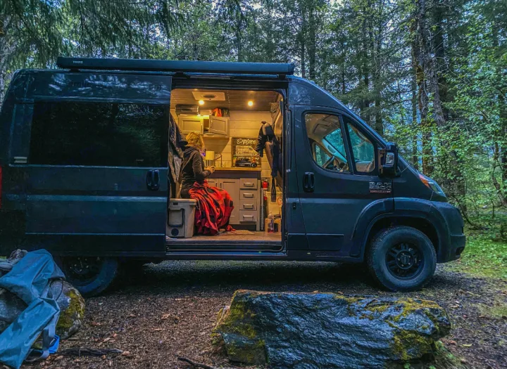 An RV rental camper van parked in a forest, its side open to reveal a cozy, lit interior with a person wrapped in a red blanket.. Ram Promaster 2020