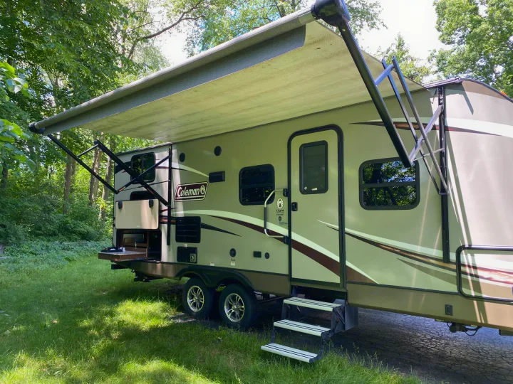 This Coleman travel trailer rv rental is set up in a wooded campsite with its awning extended and outdoor kitchen open.. Coleman Light 2405BH 2017