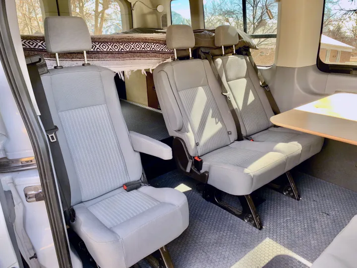 RV rental van interior featuring grey passenger seats, a bed with a patterned blanket, and a small table.. Ford Transit Custom 2019