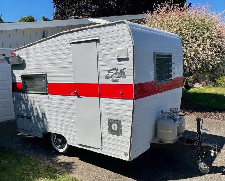Bright red and white Shasta Compact vintage travel trailer for an rv rental, parked outdoors. Classic camper with front propane tanks.. Shasta Compact 1968