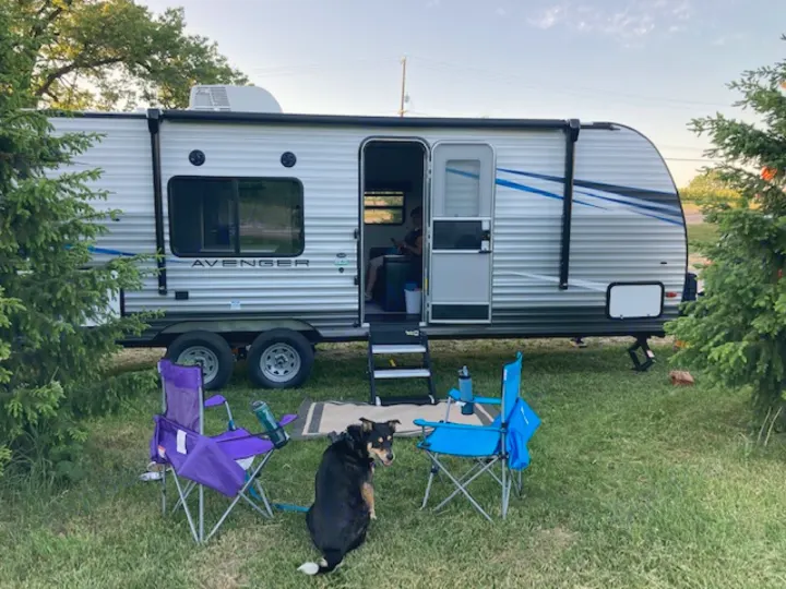 A silver Avenger RV rental is parked with purple and blue camping chairs and a dog enjoying outdoor leisure.. Forest River Prime Time Avenger 2021