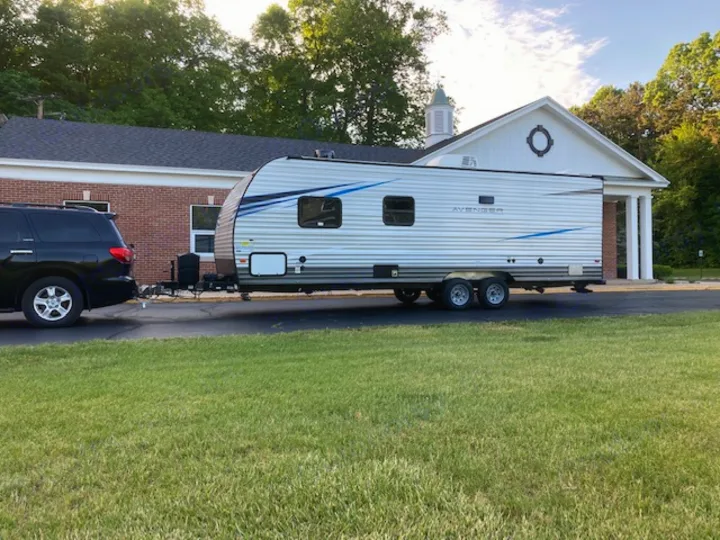 Black SUV towing a white Avenger RV rental alongside a brick building with green grass.. Forest River Prime Time Avenger 2021
