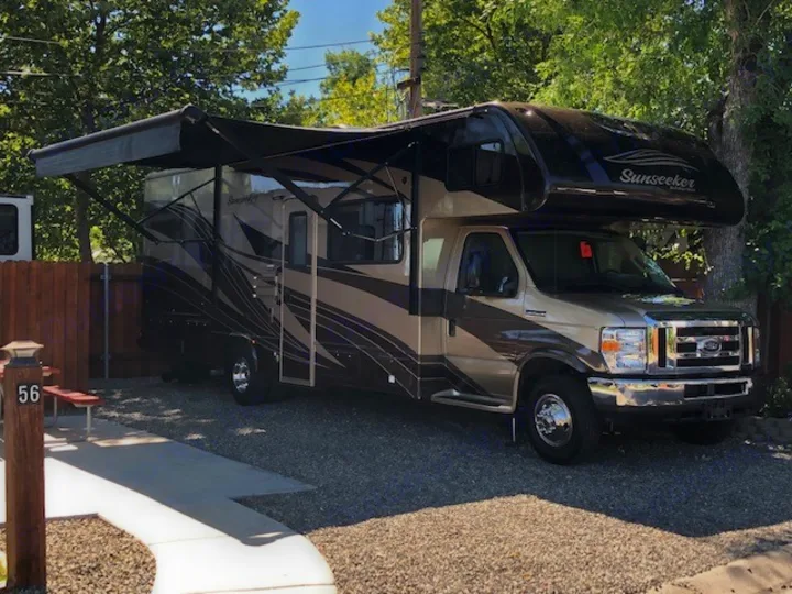 This Class C motorhome rv rental is parked at a sunny campsite with an extended awning. Brown & tan RV on gravel, site 56.. Forest River Other 2017