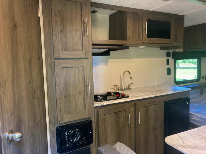 Functional RV rental kitchen with wood-look cabinets, marble-style counter, sink, cooktop, and a compact fridge.. Other 1906st Saga 2021