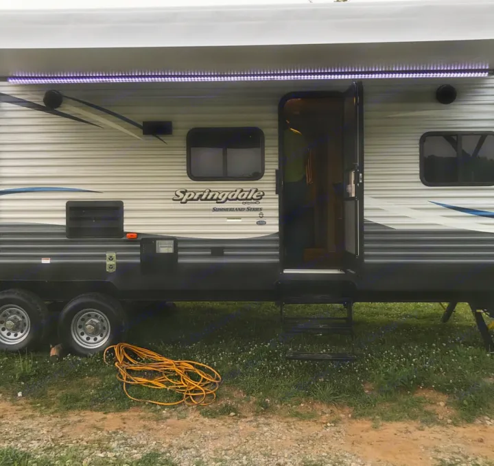 This Springdale Summerland Series RV rental shows its lighted awning, open door, and extended steps, with a yellow power cord on the ground.. Keystone Summerland 2017