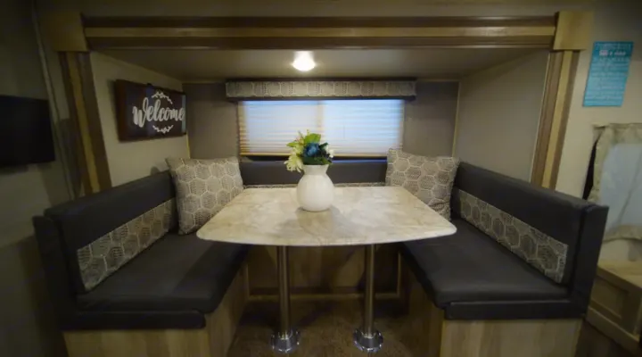 RV rental dinette: A cozy booth with a light-colored table, dark benches, patterned pillows, and a 'Welcome' sign.. Coachmen Catalina 2021