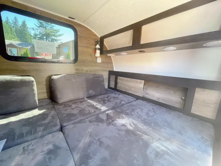 Cozy RV rental interior featuring a versatile grey sofa/bed, window with an outdoor view, and convenient overhead storage.. Braxton Creek Bushwhacker 2021