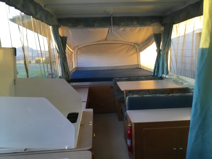Spacious pop-up RV rental interior. Features dining, kitchen, and bed areas, with outdoor views through sheer curtains.. Coleman Fairlake 2002