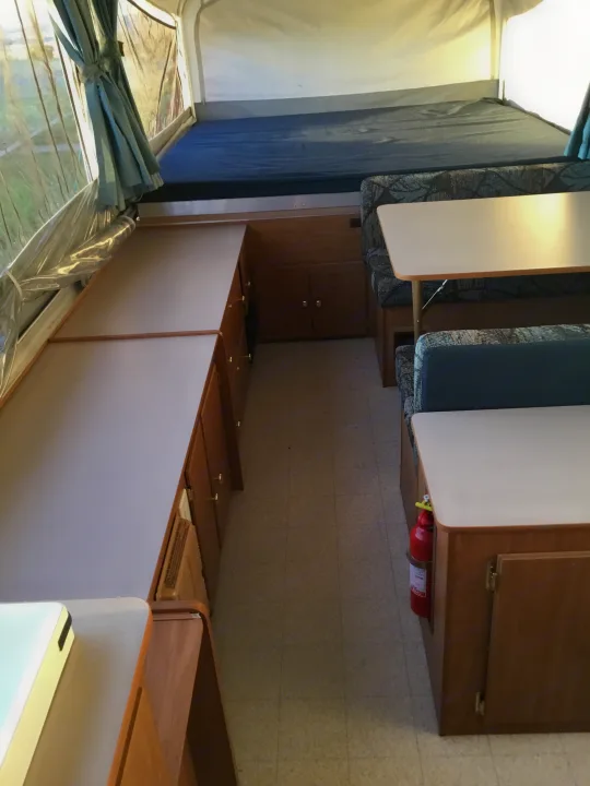 RV rental interior: A bed, dinette with blue cushions, kitchen cabinets, and a red fire extinguisher.. Coleman Fairlake 2002