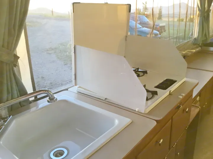 Compact RV rental kitchen: white sink & faucet, two-burner gas stove. Window reveals dirt road, trees, and hills outside.. Coleman Fairlake 2002