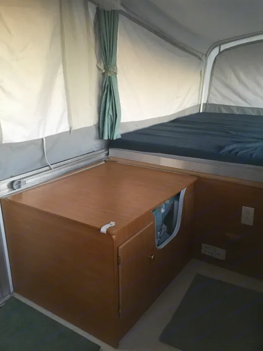 Inside an RV rental, showing a cozy pop-up bed with green drapes and a compact wooden kitchen unit.. Coleman Fairlake 2002