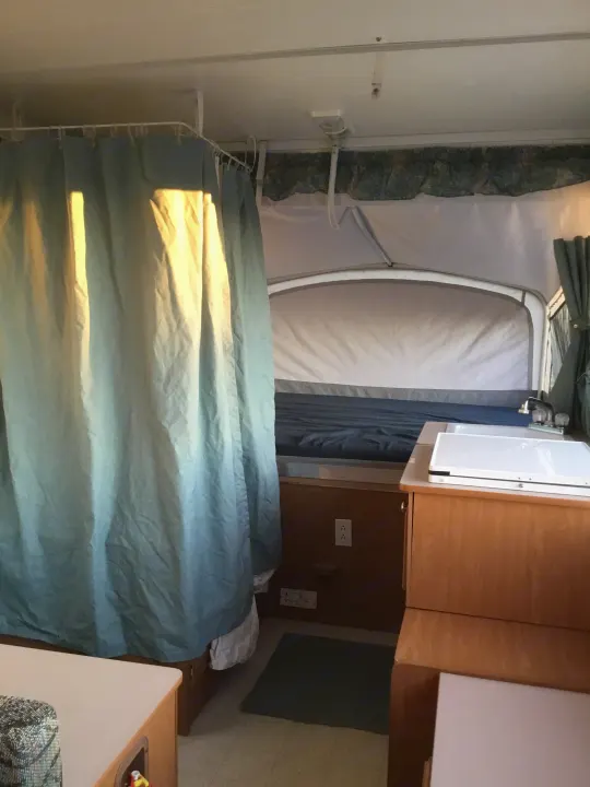Sunlit pop-up RV rental interior with bed, curtains, and wood cabinetry, offering a compact travel space.. Coleman Fairlake 2002