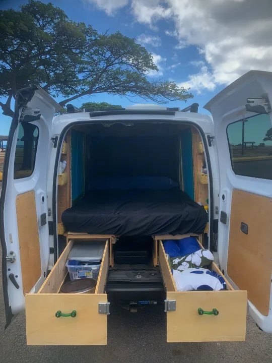 Inside an RV rental, this camper van interior features a cozy bed and custom wooden storage units with hinged panels.. Nissan NV200 2014