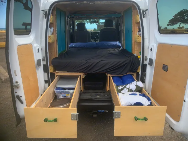 Inside a custom RV rental: a compact camper van interior featuring a cozy bed and pull-out storage, parked by the beach.. Nissan NV200 2014