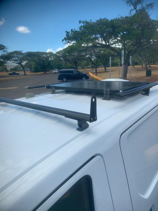 White RV rental van with a roof rack system, parked under a clear sky.. Nissan NV200 2014