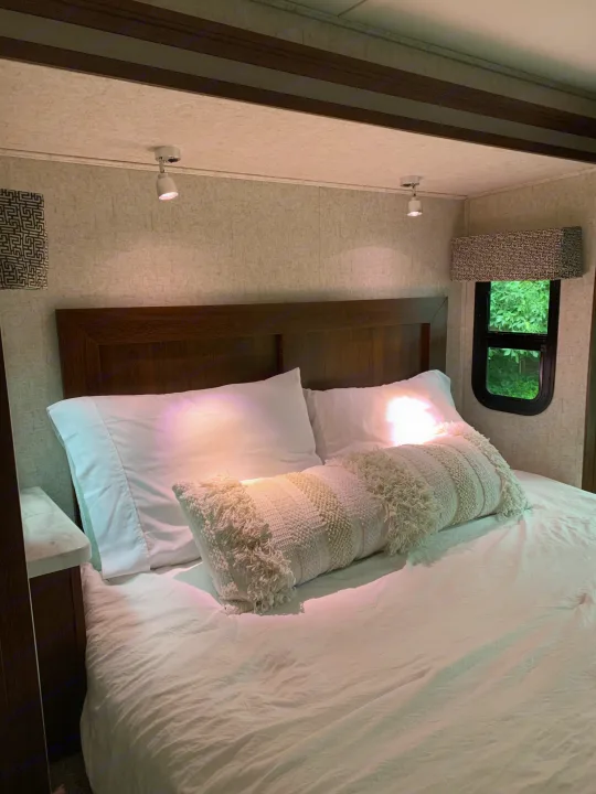 Cozy RV rental bedroom featuring a bed with white linens, a decorative long pillow, spotlights, and a window with a leafy view.. Forest River Sunseeker 2018