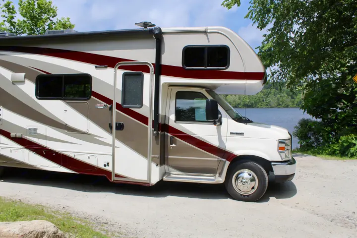 A white Class C RV rental with maroon and gray stripes is parked by a scenic lake, ideal for outdoor travel.. Forest River Sunseeker 2018