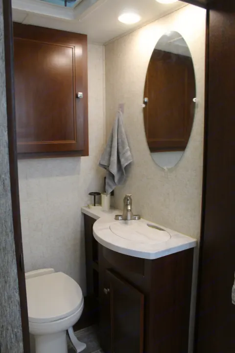 Compact RV rental bathroom showing toilet, white-topped vanity with sink, oval mirror, and dark wood cabinetry.. Forest River Sunseeker 2018