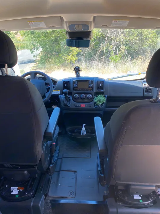 View from inside an RV rental's front cabin. Features dashboard controls, steering wheel, and front seats with a clear outdoor view.. Winnebago Other 2022