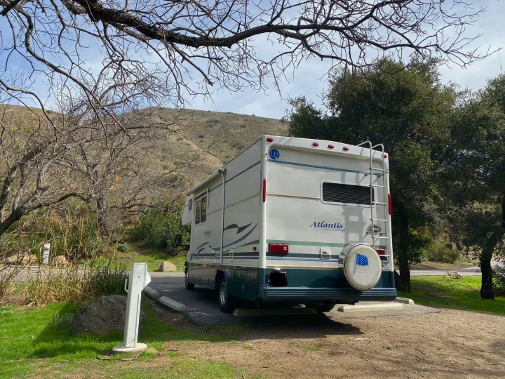 Rear view of an Atlantis RV rental parked at a sunny campsite with a utility hookup, surrounded by hills and bare trees.. Holiday Rambler Atlantis 2002