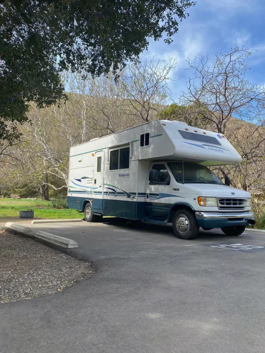 A white & teal rv rental, an Atlantis model, parked on asphalt in a campground with trees and distant hills.. Holiday Rambler Atlantis 2002