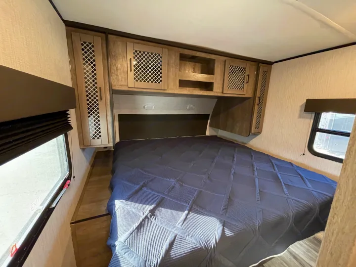 Modern RV rental bedroom features a blue quilted bed, compact overhead cabinets, and windows for a comfy travel stay.. Heartland North Trail 2021
