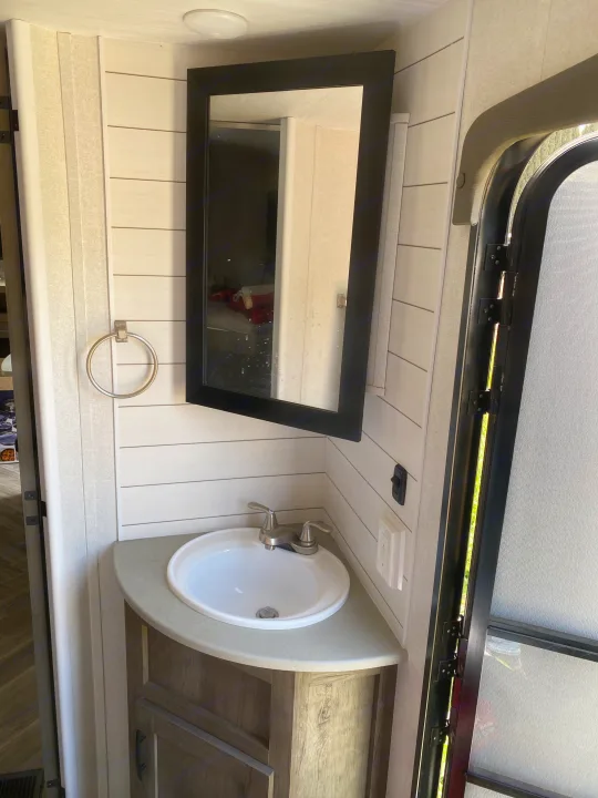 RV rental bathroom features a compact sink, black-framed mirror, white shiplap wall, and an open side door.. Forest River Evo 2020