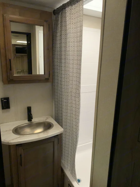 This RV rental bathroom features a vanity with a sleek sink, mounted mirror, and a curtained shower stall.. Venture Rv Sporttrek 2022