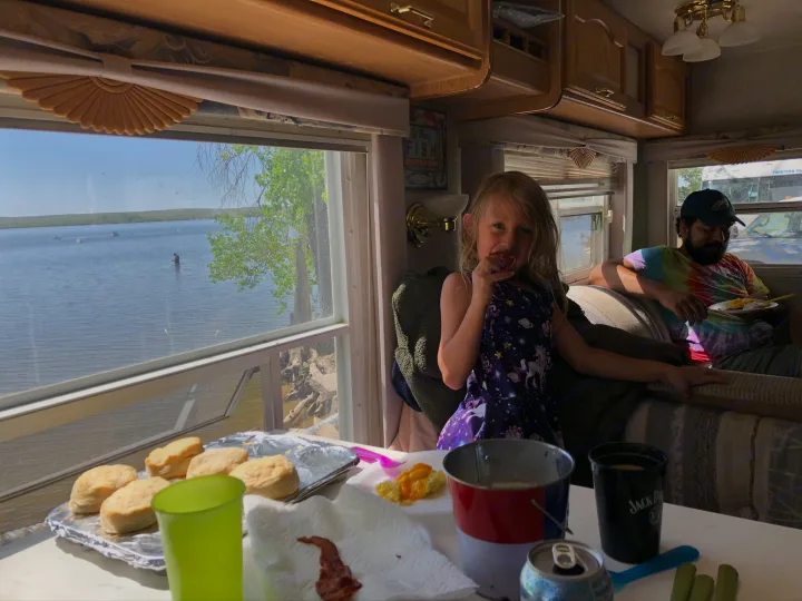 Family breakfast inside an RV rental with a scenic lake view. A girl eats at the table while a man sits nearby.. Wildcat Wildcat Trailer 2004