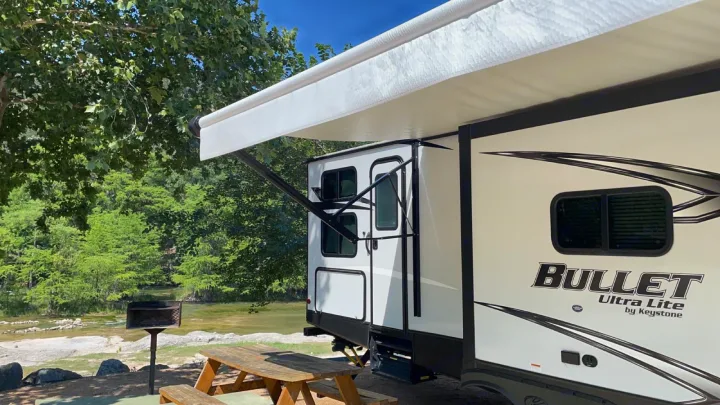 This Bullet Ultra Lite RV rental is parked by a river, featuring an extended awning, picnic table, and grill for outdoor enjoyment.. Keystone RV Bullet Ultra Lite 2021