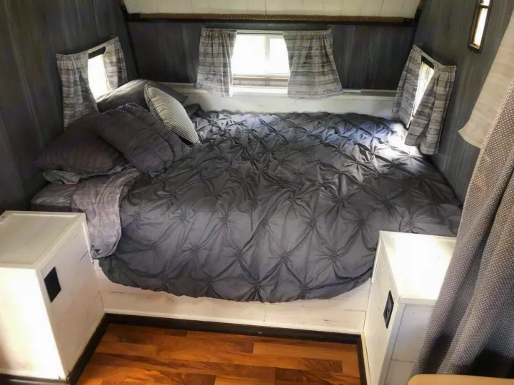 An inviting RV rental bedroom. Features a spacious gray tufted bed, multiple pillows, and side windows with curtains.. Nomad Rear fold 1962