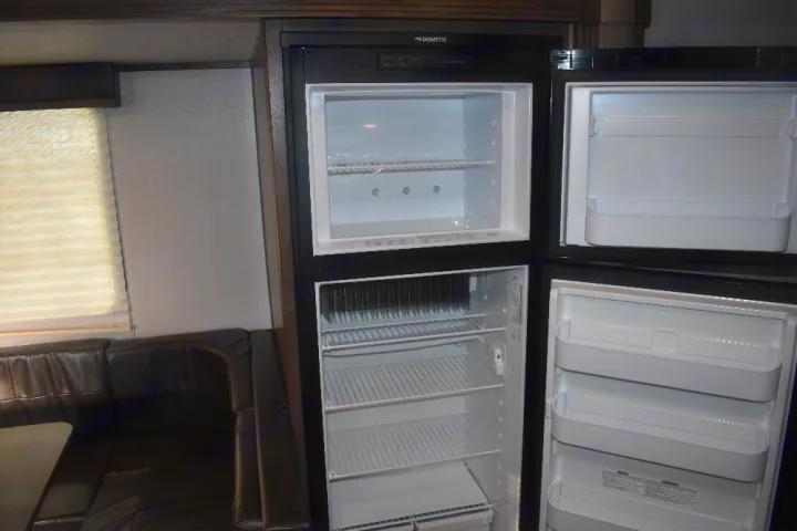 RV rental interior: Open Dometic refrigerator/freezer reveals shelves next to a dining area with a window and bench seating.. Keystone Passport 2021