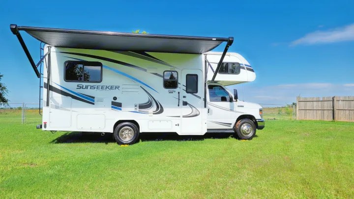 A white RV rental with an extended awning is parked on green grass under a clear blue sky, ready for travel.. Forest River Sunseeker 2019