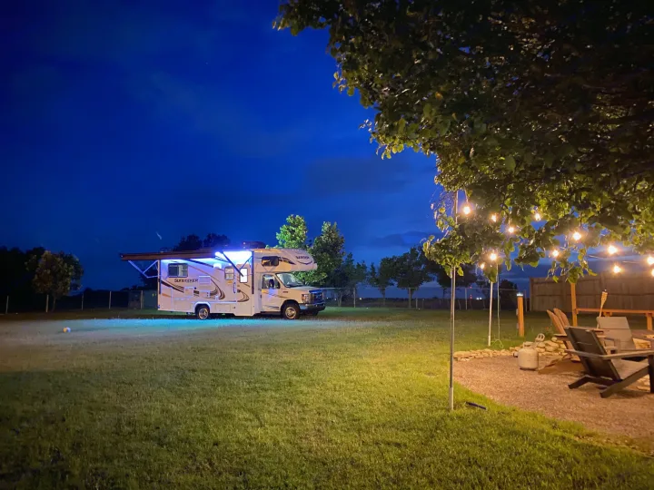 An RV rental shines with blue lights on a grassy field at night, beside a string-lit patio and fire pit for evening relaxation.. Forest River Sunseeker 2019