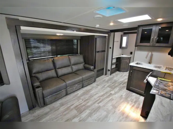 Cozy RV rental interior showcasing a gray reclining sofa, modern kitchen with sink, and bright wood-style flooring.. Grand Design Transcend Xplor 2021