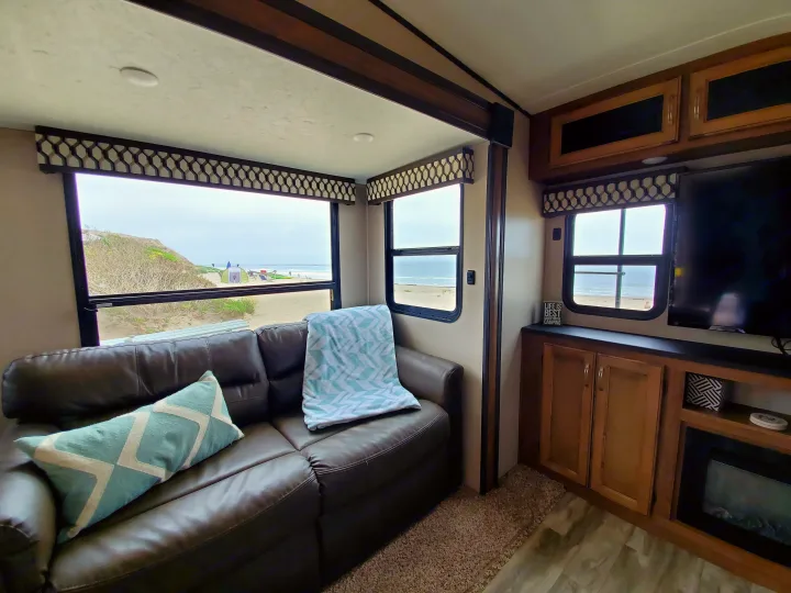 RV rental interior: Cozy living room with brown couch, TV, and large windows revealing a beautiful beach view.. Forest River Coachmen Chaparral Lite 2020