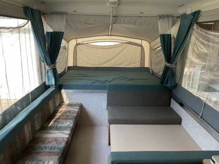 Inside a pop-up RV rental, showing a large bed, dinette, and bench seating. Teal curtains add color to this cozy space.. Coleman Destiny Cheyenne 1998
