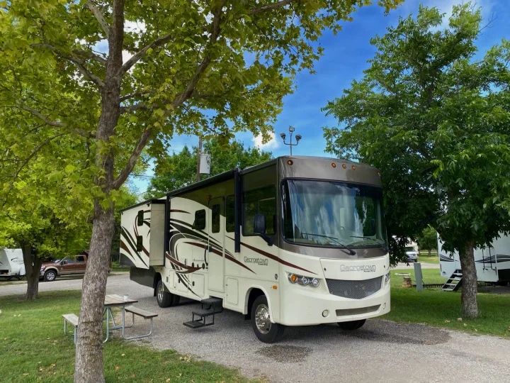 A cream and brown Georgetown motorhome RV rental, with a slide-out extended and steps down, parked at a sunny campground amid green trees.. Forest River Georgetown 2015