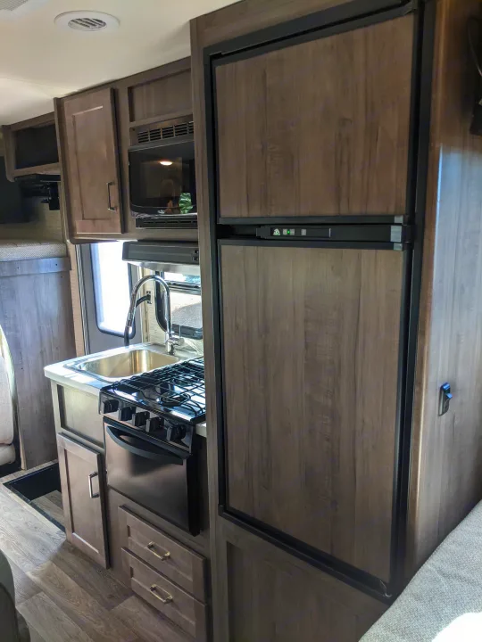 Inside an RV rental: a compact kitchen featuring a stove, sink, microwave, and refrigerator in a wood-toned interior.. Winnebago Minnie Winnie 2022